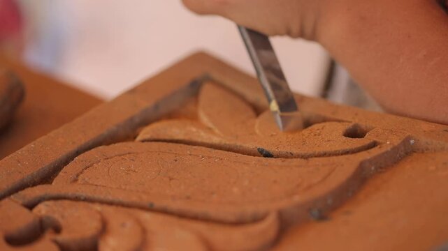 A skilled female artisan in colorful traditional attire uses a small hammer and chisel to meticulously carve an elegant floral pattern into a piece of red terracotta clay.

