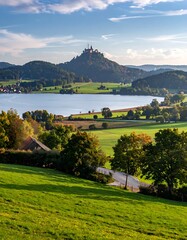 Scenic view of a castle atop a distant hill overlooking a serene lake amidst a green landscape under a cloudy sky
