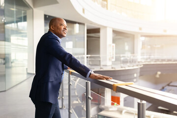 Confident mature black businessman standing in modern office atrium, leaning on railing and looking ahead with optimistic expression, corporate leadership and success concept