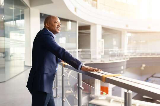 Confident mature black businessman standing in modern office atrium, leaning on railing and looking ahead with optimistic expression, corporate leadership and success concept