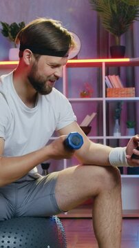Likable smiling modern active 30-aged bearded guy in white t-shirt,grey shorts and black headband sitting on rubber fitball and workouting with dumbell while revisioning intersting program on