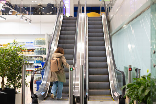 Female traveler with backpack stepping onto escalator inside airport terminal, holding boarding pass and preparing to reach departure gate