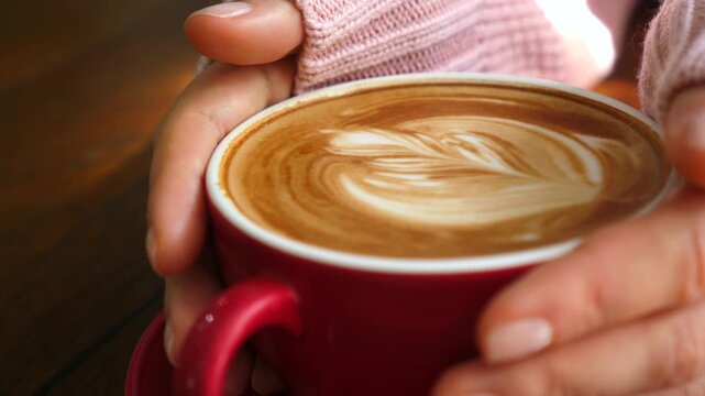 Female hands in a pink sweater gently holding a red cup of hot cappuccino with creamy rosetta latte art, evoking cozy morning coffee break and warm cafe comfort