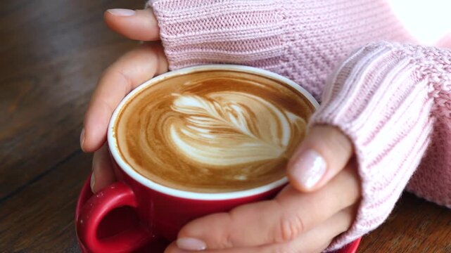 Close up of female hands in a cozy pink sweater holding a red cup of hot cappuccino with beautiful rosetta latte art on a wooden table, enjoying a warm beverage in a coffee shop