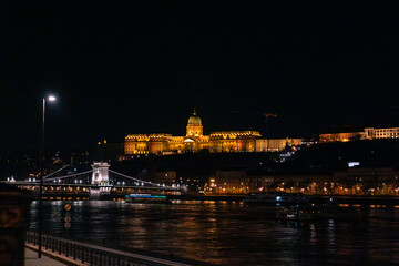 Obraz premium Buda Castle illuminated at night, viewed from the Danube River. The Chain Bridge is visible in the foreground, reflecting on the water.