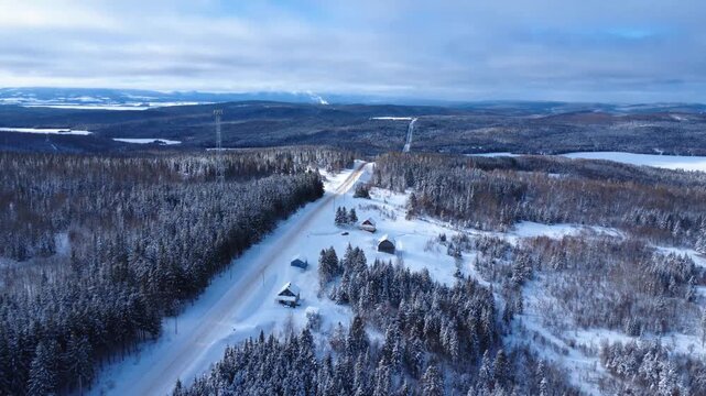Winter aerial view of a snow-covered rural road winding through a dense forest, dotted with cabins and frozen lakes, with a communications tower in a mountainous setting. Sainte-Paule, Quebec, Canada.