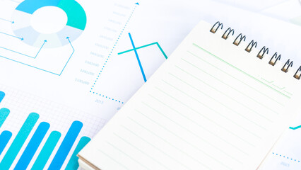 A blank notebook page sits on top of business graphs and charts with blue lines.