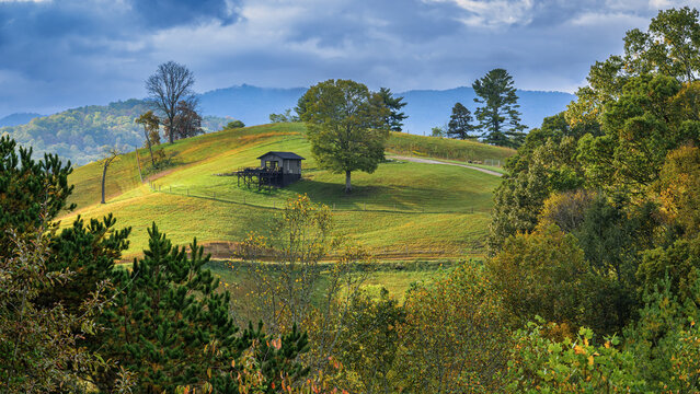 A small, dark wooden cabin with a deck sits on a grassy hillside under a cloudy sky. The surrounding landscape is dotted with trees, some showing the colors of autumn, and rolling hills recede into