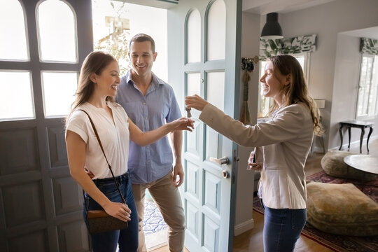 Realtor handing over house key to happy couple standing in residential doorway. Successful closing real estate, property purchase deal, home ownership, customer services, buying apartment, transaction