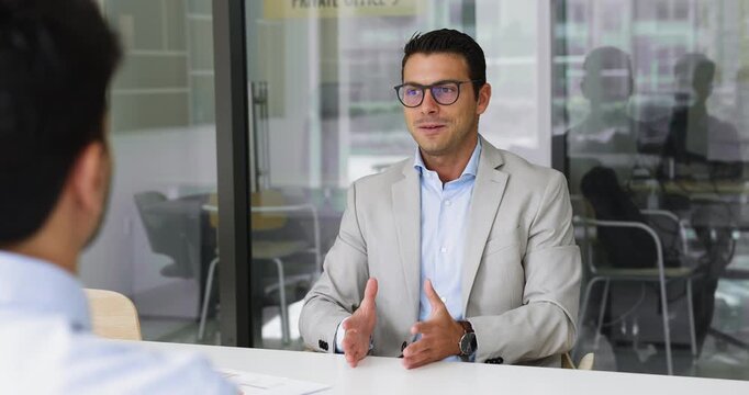 Serious, handsome young business professional man in glasses talking to HR manager on job interview, telling about soft and hard skills, sitting at meeting table, speaking