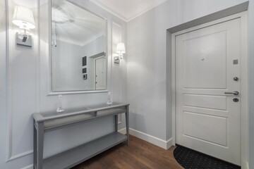 Fototapeta premium In the entrance hallway, there is a mirror on the wall above a gray table. A door leads outside. Soft light comes from the wall sconces, creating a warm feel in the area