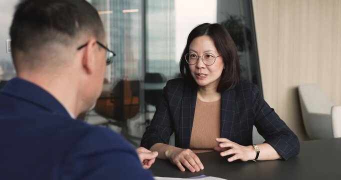 Middle-age Asian businesswoman sits at office table, shaking hands with male client or HR manager during formal meeting, job interview, as successful recruitment, business agreement and deal closing