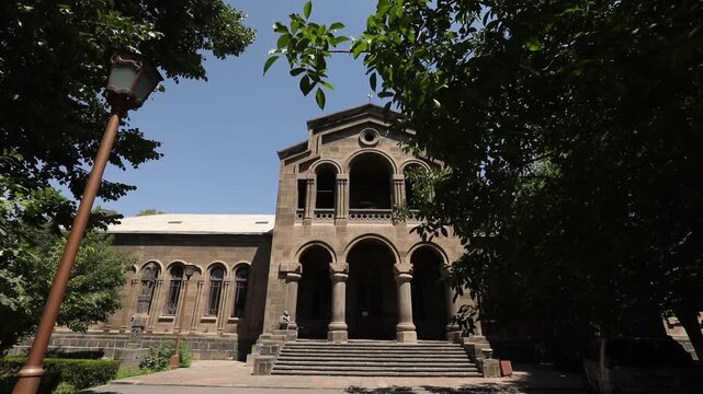 Slowly moving backward along a paved path between lush green trees to reveal the impressive facade of a historic Armenian stone building on a clear bright sunny day.