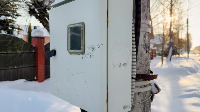 Snow covered electrical panel on pole beside cottage under soft dawn light, conduit and cables wrapped along concrete pole, rust.