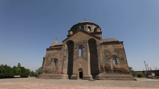 Wide shot of a historic, ancient stone church featuring a central dome and an attached bell tower, standing in a paved courtyard against a clear blue sky.

