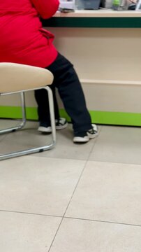 Red jacket woman waiting at reception desk, seated on chair by counter, black pants and sneakers visible, hands folded on lap.