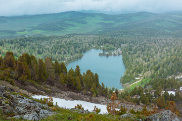 Scenic alpine top view from snowfield on scree and coniferous trees on steep slope to big wood lake against hilly vastness far away in low clouds. Long forest lake and woody hills under cloudy sky.