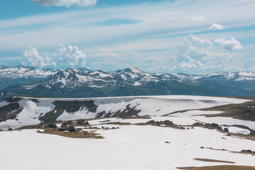 Scenic alpine landscape with sunlit snowy field on stony hill with view to big mountain range with forest and snow far away under cloudy sky. Stone outcrops among snows in sunlight in high mountains. © Daniil