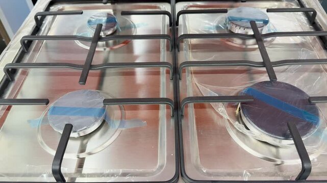 Close-up stainless gas burner blue tape showing protective packaging and polished surface, black cast iron grate framing burner.