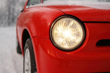 A striking close-up of a bright red car's illuminated headlight, partially covered in fresh snow. Evokes winter travel, safety, and classic automotive elegance for various campaigns.