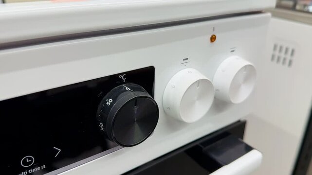 Close-up electric hob control knobs in retail showroom, glass ceramic surface reflecting aisle, white and black rotary dials.