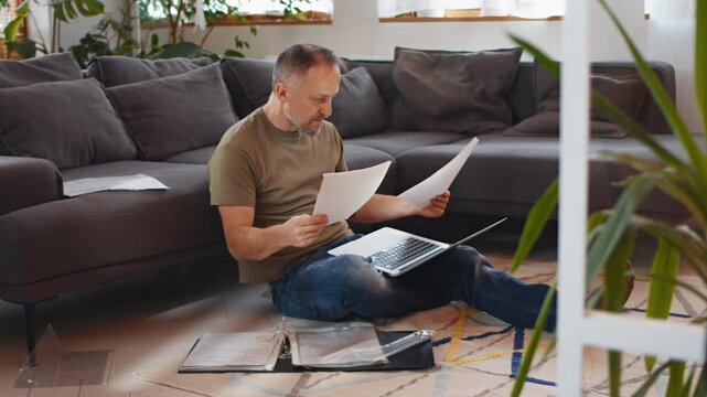 Man sitting on the floor of a living room home office and filing taxes on a laptop on his lap. Examining the paper documents in his hand