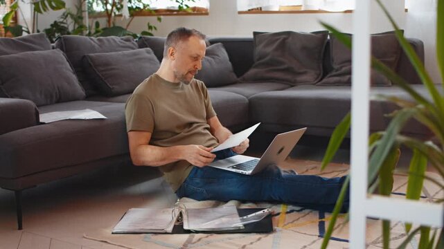 Man sitting on the floor of a living room home office and filing taxes on a laptop