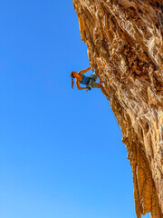 Fototapeta premium LOW ANGLE VIEW, CLOSE UP: Strong female climber on steep limestone wall with tufa formations. Young woman reaches into her chalk bag while lead climbing a steep overhang under blue Mediterranean sky.