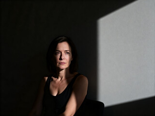 Portrait of a woman with dark hair wearing a black tank top sitting in low light with a shadow cast on the wall behind her.