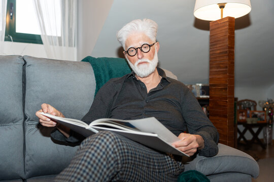 Senior Man Reading Book On Couch At Home.