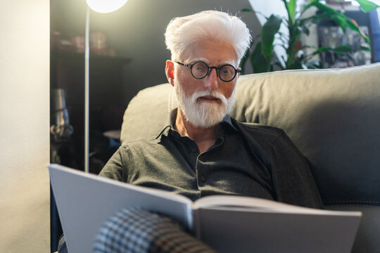 Elderly Man Reading A Book On Sofa