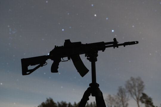 Silhouette of a modern assault rifle with holographic sight mounted on a tripod against a beautiful starry night sky background