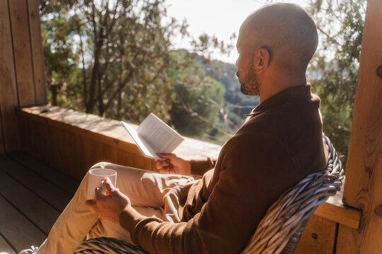 Bald Man Enjoys Tea and Book on Log Cabin Balcony in Sunny Comfort