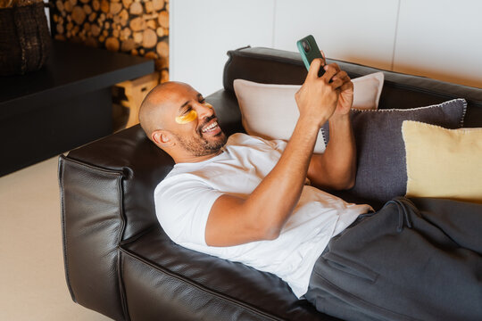 Bald Man Relaxing on Couch, Using Gel Patches and Texting at Home