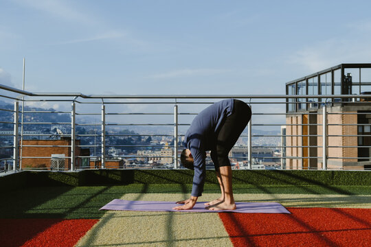 Young man doing forward bend yoga pose on rooftop with city view