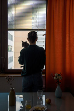 Woman holding gray cat while looking out the window