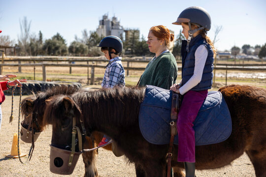 Two children in helmets sit on ponies while a woman stands beside them