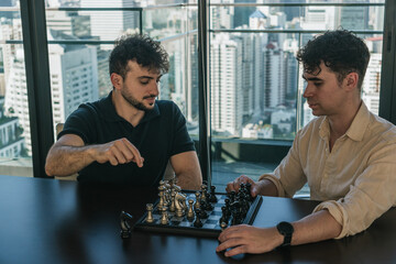 Two young men playing chess on a table by a large window with a modern city view, focusing on strategy © Felix Morion