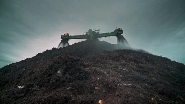 Tractor composting farm waste on a large pile under a cloudy sky