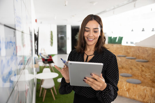 Cheerful Asian project manager man analyzing Internet marketing report on tablet, using digital gadget for product management, standing in office, holding device, smiling, laughing. Vertical shot