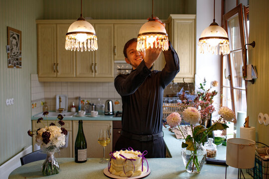 A man in the kitchen screws a lampshade into a chandelier.