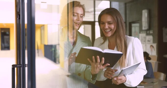 Two older adult women business colleagues reviewing information on notebook, explain or clarifying joint task or project details, reading notes, engage in team collaboration in modern coworking office