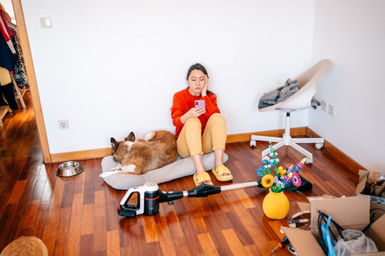 Woman sitting with dog on floor, looking at phone in messy room