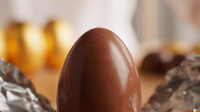 Close-up macro shot of a smooth milk chocolate Easter egg standing upright, wrapped in shiny foil, with a shallow depth of field against a subtly blurred background, evoking a festive