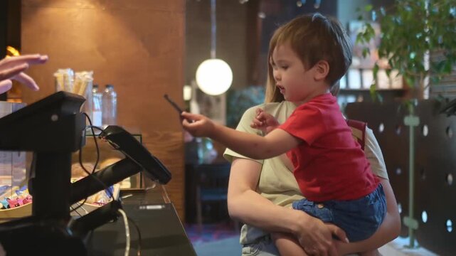 A sweet toddler boy, sitting in his mother's arms, helps her pay with a credit card at the checkout counter in a modern coffee shop. Children and money. Parents teach children how the financial system