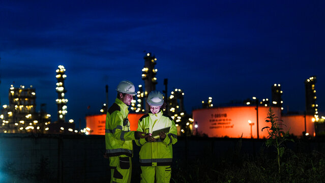 Two industrial workers in hard hats and high-vis jackets use a tablet at night. They are collaborating in front of a large, lit-up oil refinery or chemical factory plant.