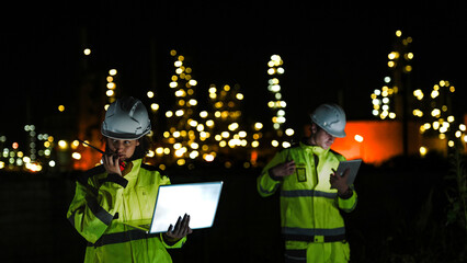 Naklejka premium Two industrial workers in hard hats and high-vis gear work at night. They use a laptop and tablet to monitor a lit-up refinery plant during a night shift.