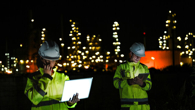 Two industrial workers in hard hats and high-vis gear work at night. They use a laptop and tablet to monitor a lit-up refinery plant during a night shift.