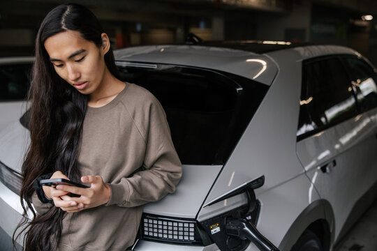 Young Woman Charging Electric Vehicle While Using Smartphone