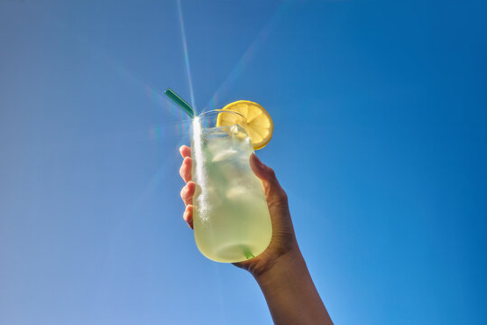 A hand holds a tall glass of lemonade with a lemon slice on blue sky